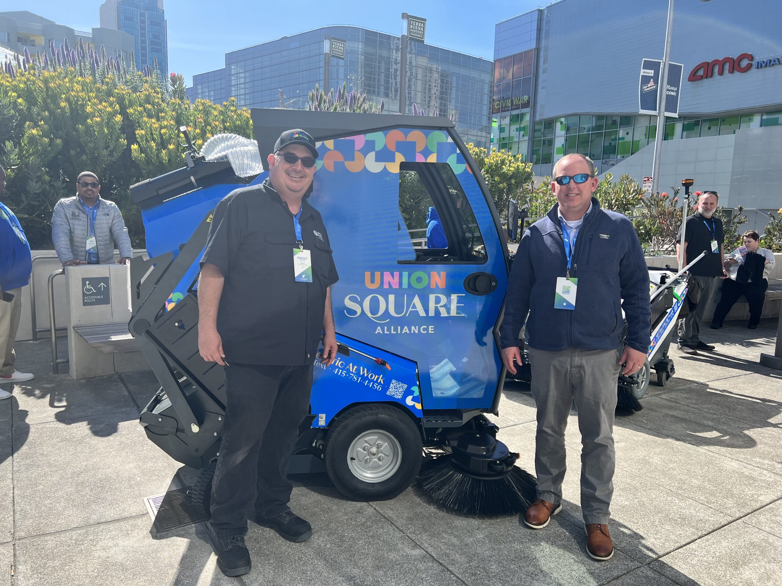 Two men stand next to a street cleaning machine called Tenax.
